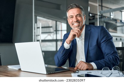 Happy smiling middle aged professional business man company executive ceo manager wearing blue suit sitting at desk in office working on laptop computer laughing at workplace. Portrait. - Shutterstock ID 2302831097