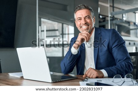 Happy smiling middle aged professional business man company executive ceo manager wearing blue suit sitting at desk in office working on laptop computer laughing at workplace. Portrait.