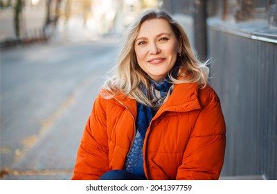 Happy lächelnde Frau mittleren Alters in orangefarbener Daunenjacke auf Beton-Treppen im Freien und mit Blick auf Kamera mit positivem Gesichtsausdruck, ruhen beim kalten Herbsttag in der Stadt