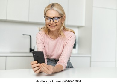 Happy Smiling Middle-aged Woman Holds Smartphone Sitting In The Kitchen. Charming Lady Spends Leisure Time Websurfing, Shopping Online, Scrolling News Feed On The Phone At Home