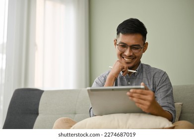 Happy and smiling millennial Asian man in casual clothes using his digital tablet, watching video or working on his project, sitting on sofa in his living room. - Powered by Shutterstock