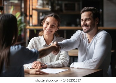 Happy smiling millennial couple handshake get acquainted with female real estate agent meeting together in cafe, excited husband and wife shake hand of broker or banker thanking for help