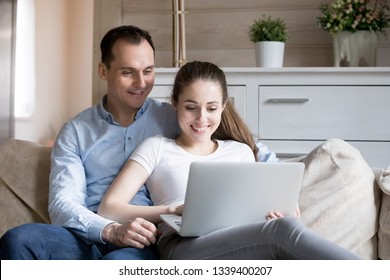 Happy Smiling Millennial Couple Watching Video On Computer At Home. Man And Woman Sitting On Sofa Looking At Laptop Screen Having Fun On Weekend Together. Family Leisure Time. Gadget Addiction Concept