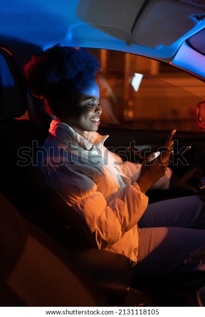 Happy smiling millennial girl sit in car\
scrolling social media after work. Young african female smartphone\
addict use mobile phone sitting alone in vehicle at dark empty\
parking. Gadget addiction