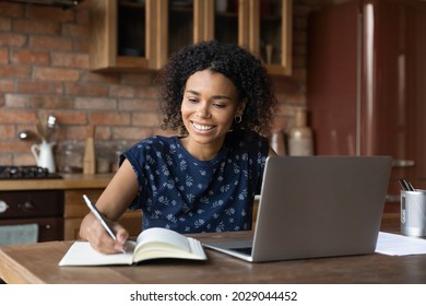 Happy smiling mixed race African student girl studying from home, writing notes, watching, listening, summarizing learning webinar on laptop, attending virtual school class, online college lesson - Powered by Shutterstock