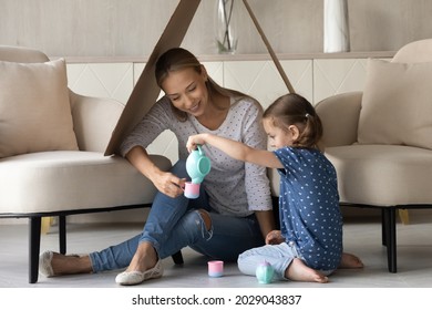 Happy Smiling Mom And Little Daughter Kid Playing Tea Party In Toy Handmade House, Sitting On Heating Floor Under Cardboard Roof, Using Colorful Plastic Dish For Kids. Childhood, Housing, Imagination