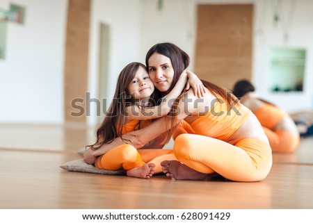 Happy smiling mother and daughter in bright orange sport suits sitting on the floor in the gym, hugging, looking at the camera