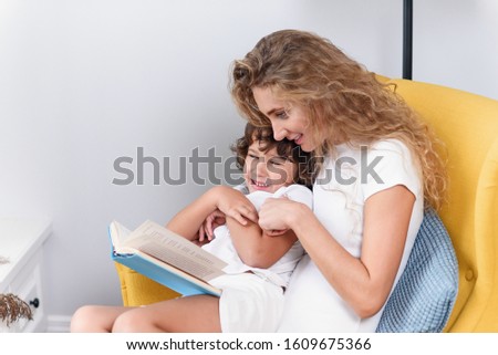 Happy smiling mother and her funny son in white clothes having fun together while reading interesting educational book.