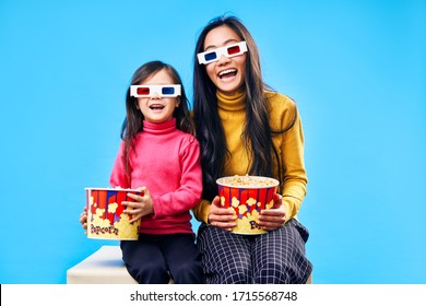 Happy Smiling Mother And Her Little Daughter In 3D Glasses With Popcorn Watching Movie Over Blue Background