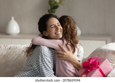 Happy smiling mother hugging thanking little daughter, holding wrapped pink gift box, sitting on couch at home, adorable cute girl kid congratulating excited mom with birthday or mothers day