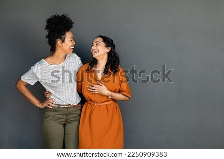 Happy smiling multiethnic women embracing each other against grey wall with copy space. Happy laughing girls standing on grey background and looking at each other. Carefree girls having fun.