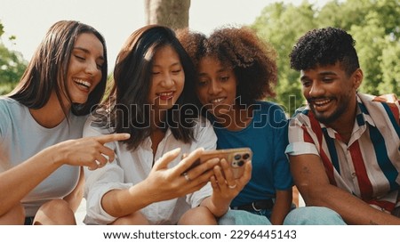 Happy, smiling multiethnic young people at picnic on summer day outdoors. Group of friends talking, using cellphone while relaxing in the park at picnic