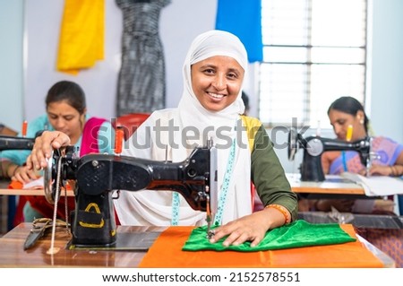 Happy smiling muslim woman in hijab loooking at camera while stitching using sawing machine at garments - concept of equality, diversity and employment