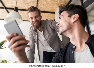 Happy smiling office worker is holding phone. His merry colleague standing near him and looking at smart phone. Low angle