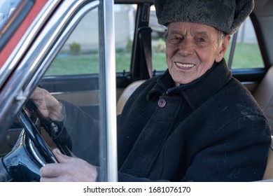 Happy Smiling Old Man In Black Jacket Driving Retro Car, Holds Her Hands On Wheel And Looking At The Camera, Outdoor Photo