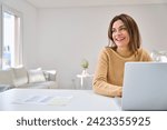 Happy smiling older mature middle aged woman using laptop computer sitting at table in living room, hybrid working, elearning online with technology device at home, looking away at copy space.