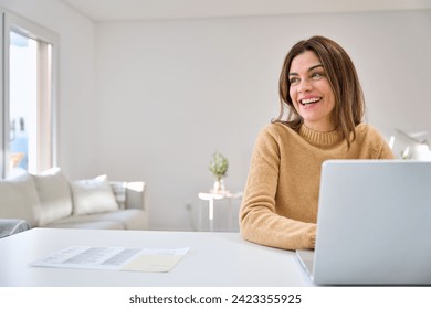 Happy smiling older mature middle aged woman using laptop computer sitting at table in living room, hybrid working, elearning online with technology device at home, looking away at copy space. - Powered by Shutterstock