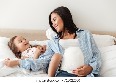 Happy smiling pregnant asian woman having fun with her daughter while sitting on bed together