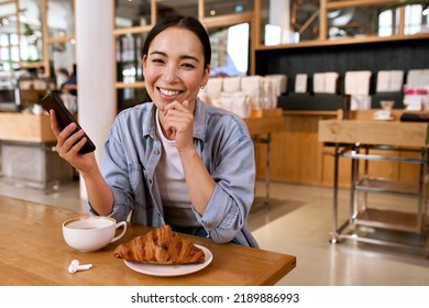 Happy smiling pretty young Asian woman user student holding smartphone device using dating application on cellphone, laughing looking at camera, having fun sitting at cafe table enjoying coffee. - Powered by Shutterstock