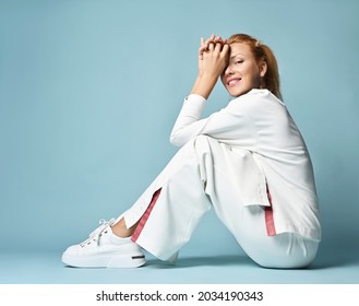 Happy smiling red-haired woman citizen in white trendy casual pantsuit with sweater, kimono style and white sneakers sits sideways on floor holding hands at head and looks at camera