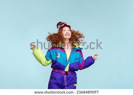 Happy smiling redhead curly woman wearing stylish ski overall, jumping looking away isolated on blue background. Concept of shopping, advertisement