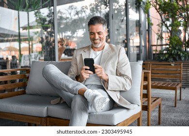 Happy smiling relaxed mid aged business man, mature professional businessman entrepreneur sitting in outdoor cafe holding smartphone using mobile phone digital technology apps. Authentic shot - Powered by Shutterstock