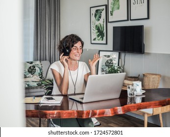 happy smiling remote online working woman with laptop, headset, notebook, mobile, pen and glass in casual outfit sitting on a work desk in her living room in her home office having video chat