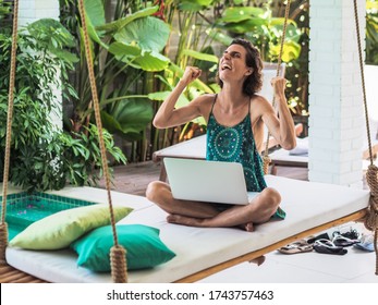 happy and smiling remote online working digital nomad women in winner pose with a laptop sitting at on a swing bed next to a turquoise water pool with beach chairs and plants in the background