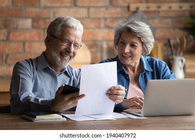 Happy smiling retired family couple do paperwork at home office at kitchen in house pay utility bills online using laptop calculate expenses. Friendly elderly spouses check finance papers plan budget
