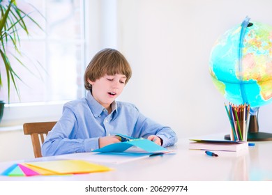 Happy Smiling School Boy, Smart Student, Doing Homework Cutting Paper, Writing, Drawing And Reading A Book At A White Desk With A Globe Next To A Window, Back To School Concept