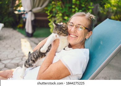 Happy Smiling Senior Elderly Woman In Glasses Relaxing In Summer Garden Outdoors Hugging Domestic Tabby Cat. Retired Old People And Animals Pets Concept