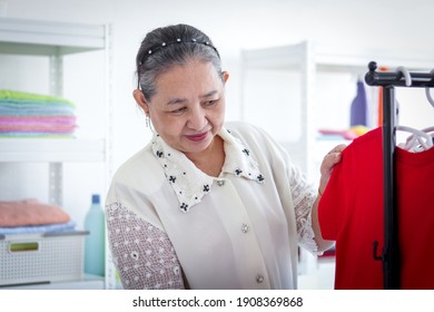 Happy Smiling Senior Elderly Woman Housewife Hanging Clothes Indoor To Dry, Older Asian Female Doing Laundry At Laundry Room, Dried Laundry, Grandma Doing Housework And Cleaning Clothes At House.