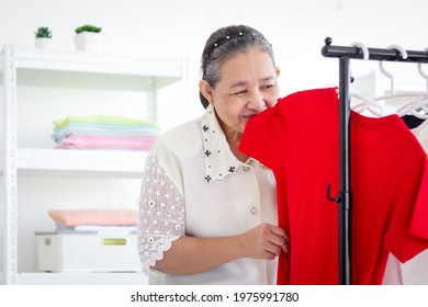 Happy Smiling Senior Elderly Woman Housewife Smelling T-shirt, Sniffing From Clothes, Hanging Clothe,  Older Asian Female Doing Laundry, Grandma Doing Housework And Cleaning Clothes At House.