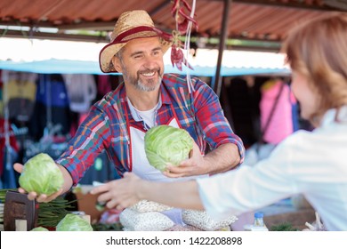 Happy Smiling Senior Farmer Standing Behind The Stall, Selling Organic Vegetables In A Marketplace. Serving Mature Female Customer. Healthy Organic Local Food Production