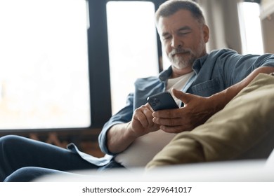 Happy smiling senior man using smartphone device while sitting on sofa at home. Mature man lying on couch reading messages on mobile phone, relaxing at home. - Powered by Shutterstock