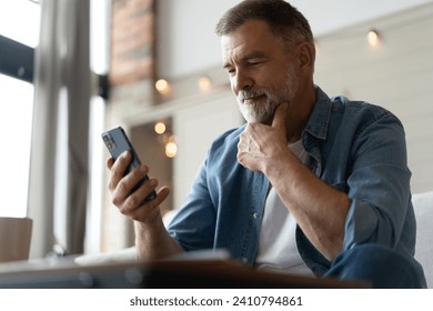 Happy smiling senior man using smartphone device while sitting on sofa at home. Mature man lying on couch reading messages on mobile phone, relaxing at home. - Powered by Shutterstock