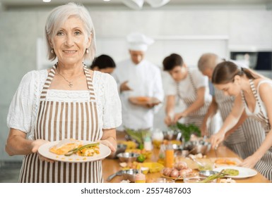 Happy smiling senior woman participating in group cooking class, standing in striped apron with plate of prepared dish in hands.. - Powered by Shutterstock