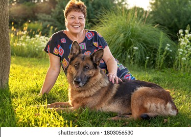 Happy smiling senior woman sitting on grass with German Shepherd dog in garden outdoors at sunset. Family pet. Friendship and care - Powered by Shutterstock