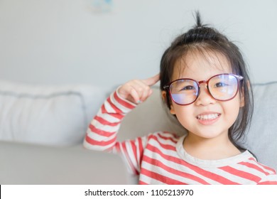 Happy Smiling Smart And Nerd Little Asian Girl In Glasses And Pointing Finger At Brain Holding Laptop In Sofa Living Room.Child With School Kindergarten.Child Development And Education Concept.