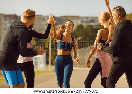 Happy smiling sporty young friends giving high five to a woman rejoicing the success in sport training after jogging exercises outdoors. People standing in the park after workout in nature.