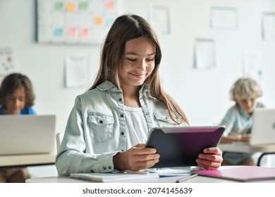 Happy smiling teen elementary schoolgirl studying looking at tablet device sitting in classroom with group of schoolchildren using laptop computers. Modern technologies for education concept.