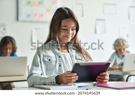 Happy smiling teen elementary schoolgirl studying looking at tablet device sitting in classroom with group of schoolchildren using laptop computers. Modern technologies for education concept.