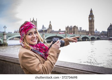 Happy, smiling tourist woman in beige coat sightseeing and points to Big Ben with pink shawl and camera - England, London, UK