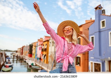 Happy smiling traveler woman posing in street, among colorful houses on Burano island. Girl wearing trendy summer outfit with orange sunglasses, straw hat, pink shirt. Copy, empty space for text
