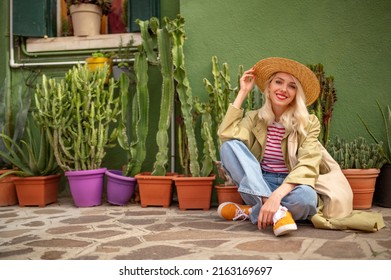 Happy smiling traveler woman wearing trendy spring, summer outfit with straw hat posing, sitting near green color house with many plants. Travel, tourism, lifestyle concept. Copy, empty space for text
