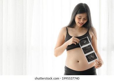 Happy smiling two months Asian pregnant woman holding ultrasound scan images by the window 