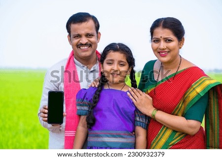 Happy smiling village couple with kid showing green screen mobile phone by pointing finger while looking camera at farmland - concept of advertisement, app promotion and family bonding.