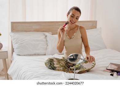 Happy smiling vivid young woman in casual clothes sit in bed look in small mirror doing make up rest relax spend time in bedroom lounge home in own room house wake up dream be lost in reverie good day