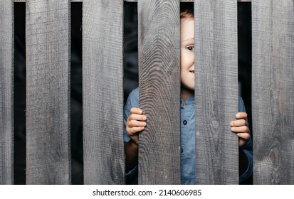 Happy Smiling White Boy Looks Out Of The Crack Of A Wooden Fence. Childish Curiosity. Espionage. Rural Life. Child Development, Curiosity. The Boy Peered Through The Crack.