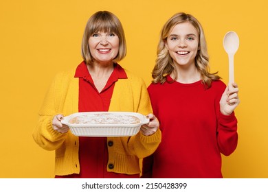 Happy Smiling Woman 50s In Red Shirt Have Fun With Teenager Girl 12-13 Years Old. Grandmother Granddaughter Hold Spoon Meal Dish Casserole Isolated On Plain Yellow Background. Family Lifestyle Concept